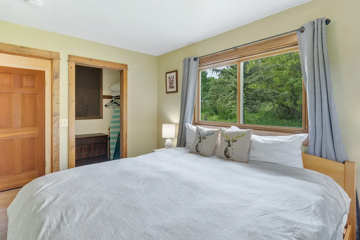A serene bedroom featuring a plush bed with decorative pillows, a large window offering scenic views, and a nightstand with a lamp. The space is bathed in natural light, creating a tranquil setting for relaxation.