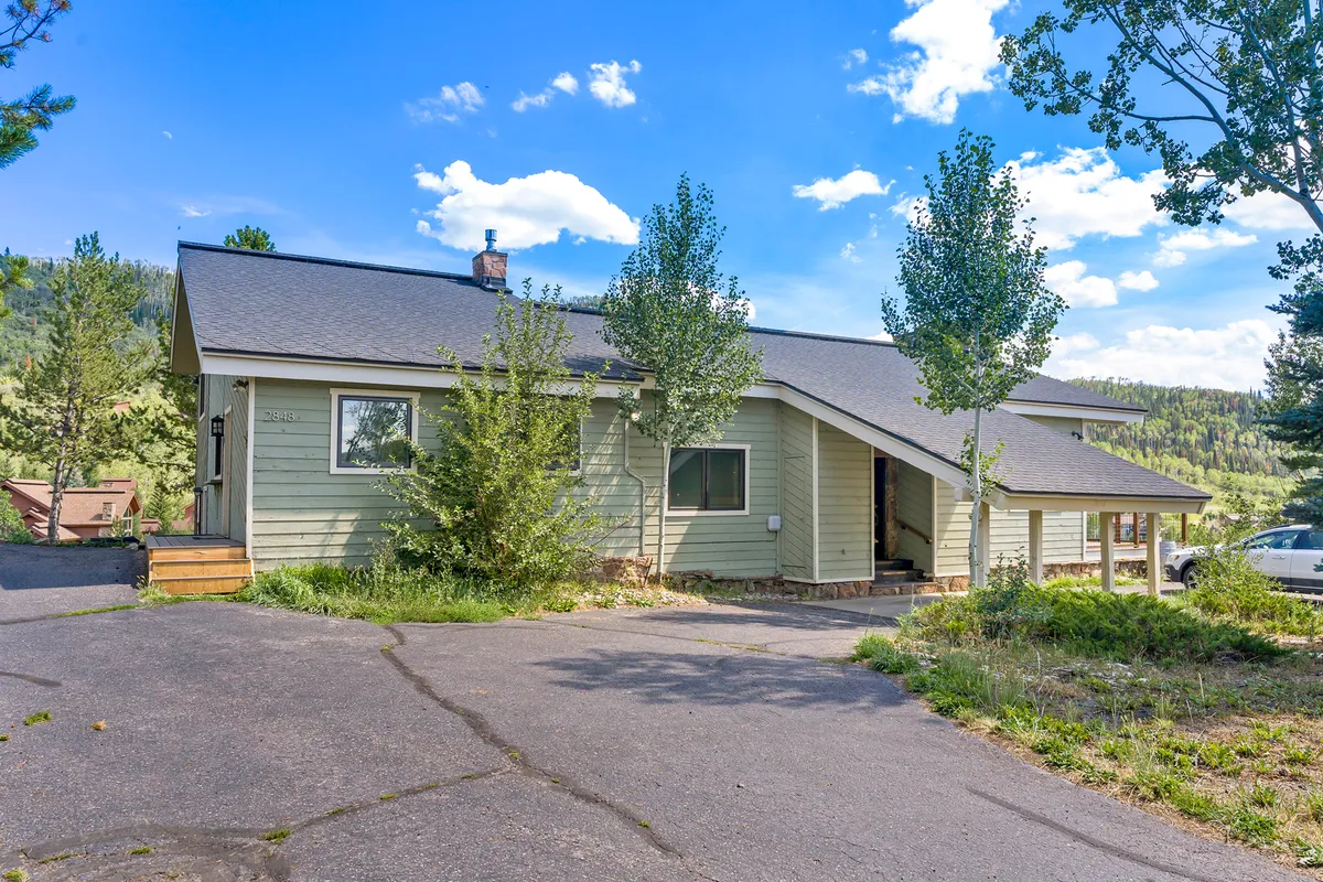 The image showcases the exterior of a cozy single-family house with an angled roof and large windows, nestled amid lush greenery. The house is surrounded by trees, and the porch offers inviting views, perfect for enjoying a sunset or morning coffee in a tranquil mountain setting.
