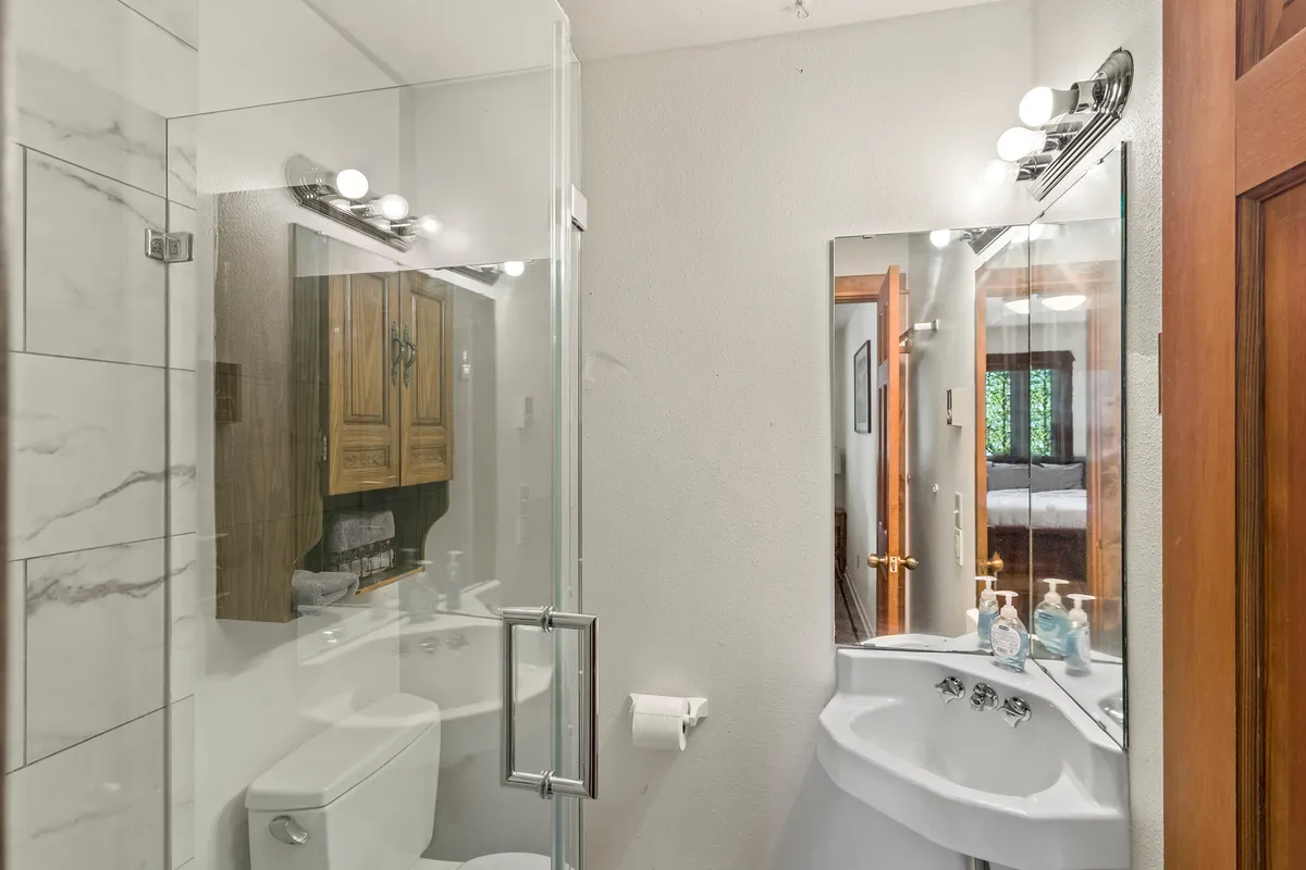 A modern bathroom with a glass shower enclosure, a wooden vanity with a framed mirror, and sunlight streaming through the window. The scene feels fresh, clean, and inviting, ideal for morning routines or unwinding after a day out.