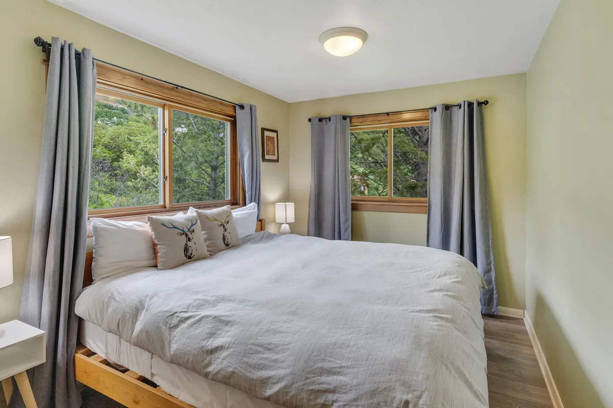 A comfortable bedroom with a large bed positioned near windows that offer a view of the outdoors. Natural light filters through the curtains, creating a bright and inviting space ideal for relaxing after exploring mountain trails.