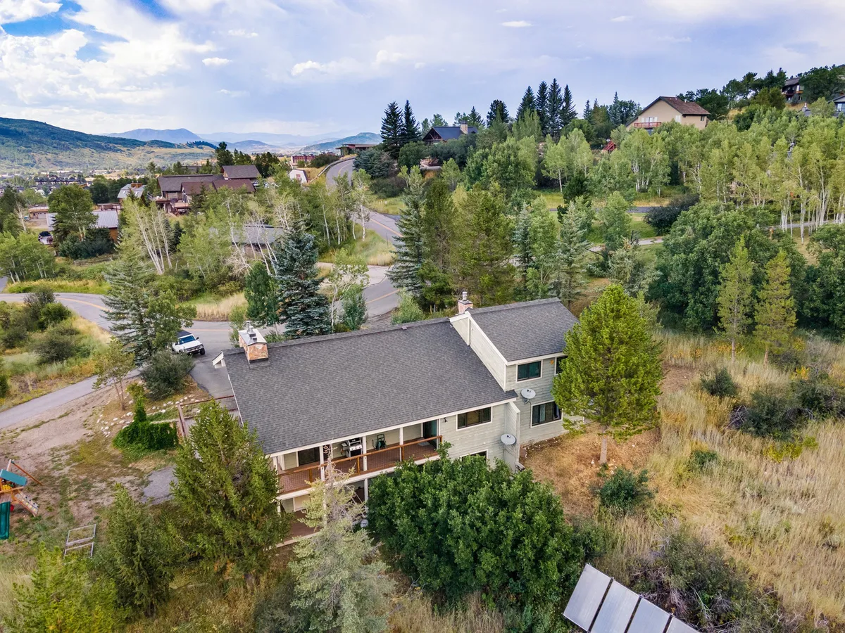 A view of the duplex exterior surrounded by trees, with a nearby road visible. The house features an inviting entrance and a tranquil natural setting, hinting at proximity to outdoor activities and mountain adventures.