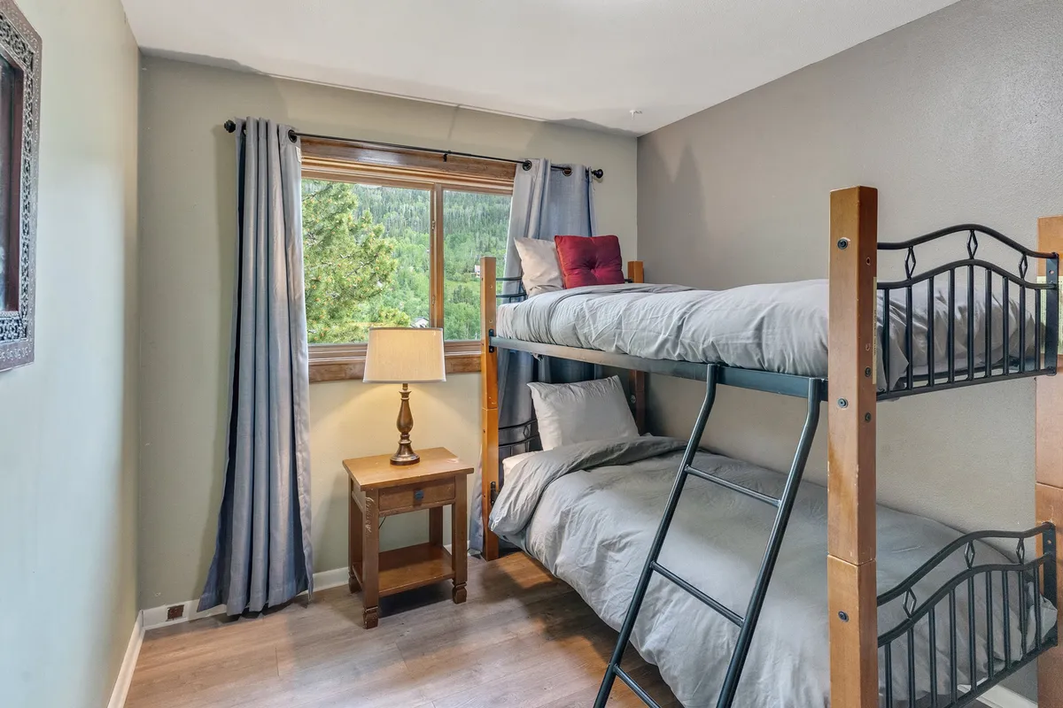 A bright and cheerful bunk room with cozy bunk beds, a window view, a nightstand with a lamp, and curtains. The setup is playful yet tidy, ideal for kids or groups wanting a fun sleepover environment.