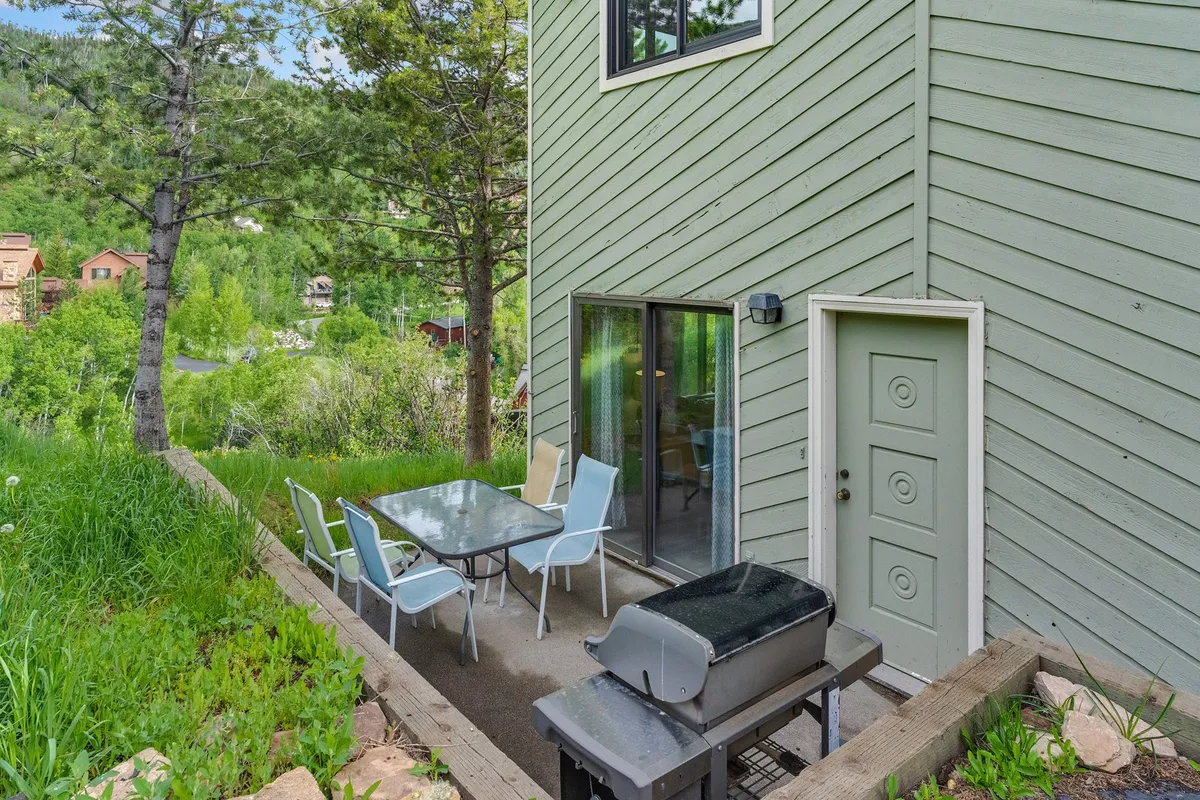 This image features a private patio area complete with a patio table, BBQ grill, comfortable seating, and lush green landscaping. The setting is framed by sliding glass doors, seamlessly connecting the indoor living space with the outdoors. The tranquil atmosphere and well-appointed outdoor furniture invite guests to relax, dine, or enjoy a sunset meal with friends, family, and pets.