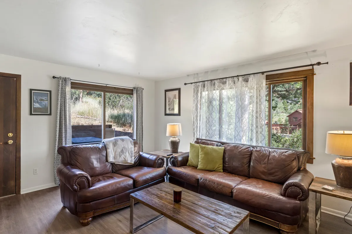 A welcoming living space featuring leather sofas and a wooden coffee table. Large windows allow daylight to fill the room, and a cozy lamp adds ambient lighting, creating an inviting spot for relaxing or socializing.